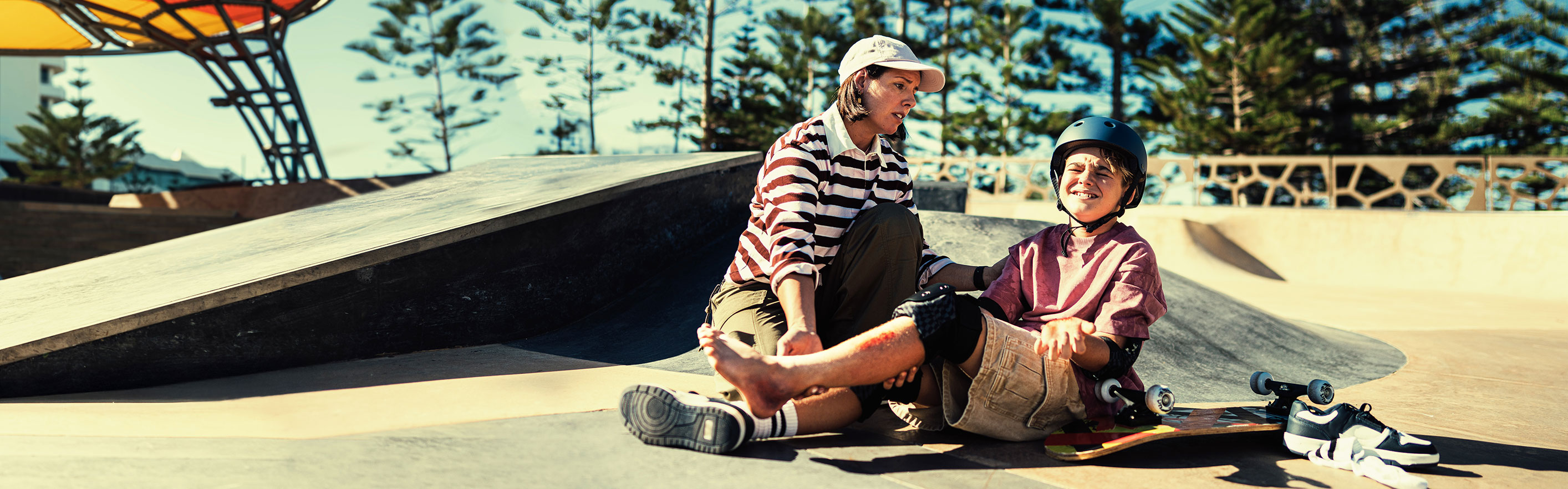 Young mother kneeling helping young son with an ankle injury at a skatepark.