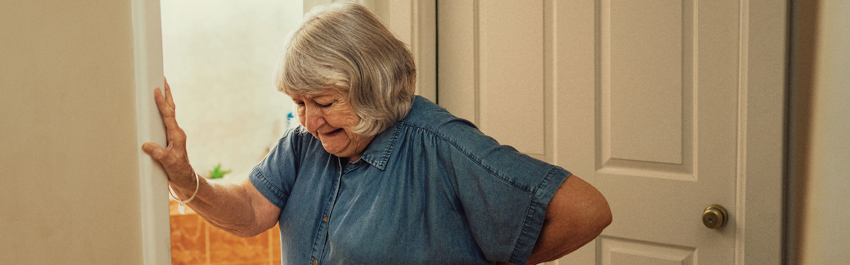 Older woman leaning against a wall in a hallway, holding lower back due to UTI pain